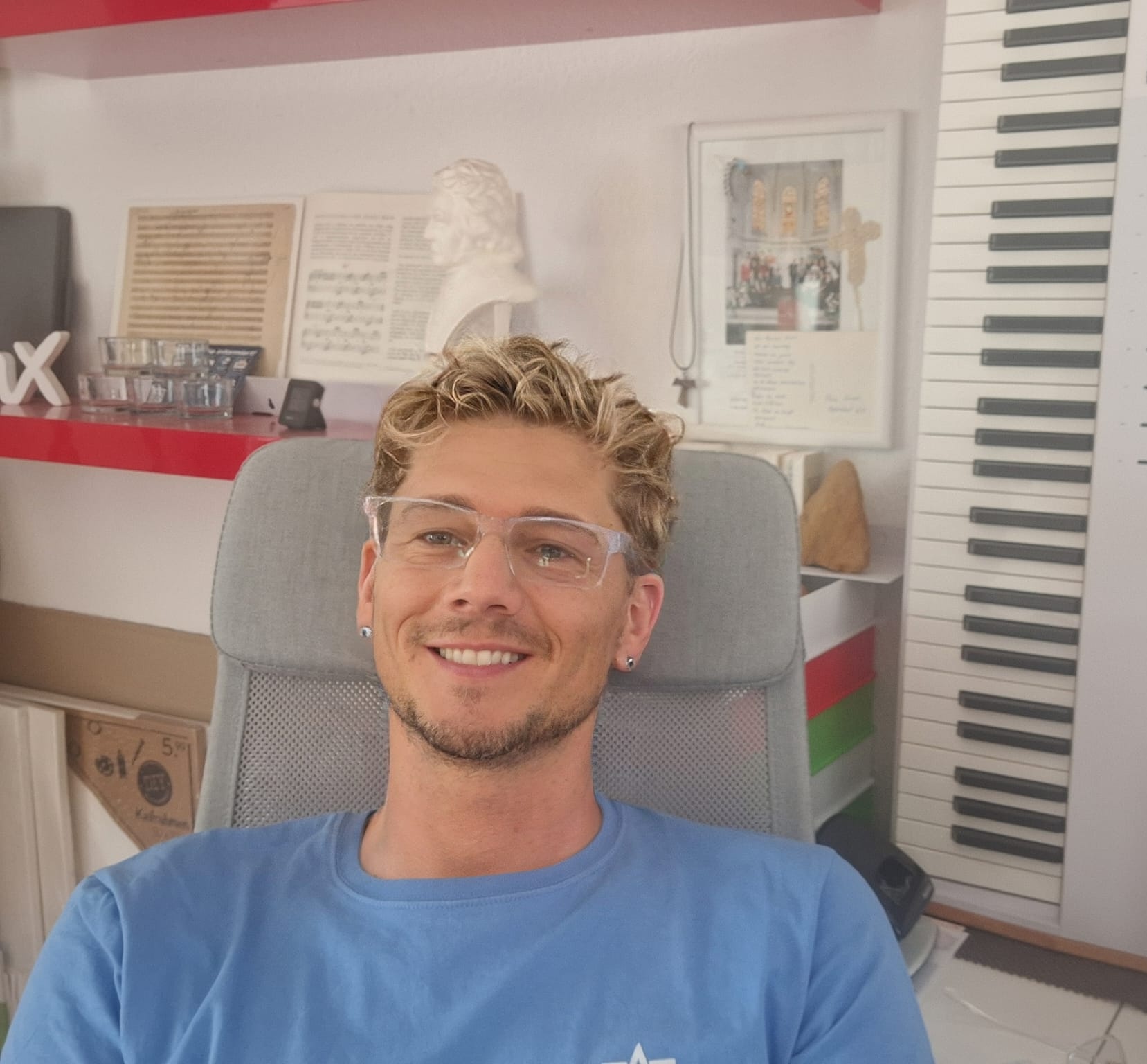 Gründer und Leiter des Schulchors David Sindt Die Grafik zeigt im oberen Bereich ein Porträt von David Sindt, dem Gründer und musikalischen Leiter des Geschwister Scholl Chors, sitzend, vor einem hochkant an die Wand gelehrten Masterkeyboard. Im Hintergrund sind eine weiße Beethoven-Büste vor einem historischen Noten-Faksimile von „Für Elise“ zu sehen, während darunter ein gedrucktes Zitat des Pianisten Jan Lisiecki über die Zugänglichkeit und den Anspruch klassischer Musik zu lesen ist. • Es lautet: „Ich lehne die Vorstellung ab, dass klassische Musik elitär ist. Ich mag es, sie für alle zugänglich zu machen, jung und alt, in großen und kleinen Städten. Auf der anderen Seite gefällt es mir überhaupt nicht, wenn Leute versuchen, das Niveau zu senken, um es vermeintlich verständlicher zu machen.“ • David Sindt kommentiert wie folgt: „Ein Zitat des Pianisten Jan Lisiecki, dem nichts hinzuzufügen ist.“
